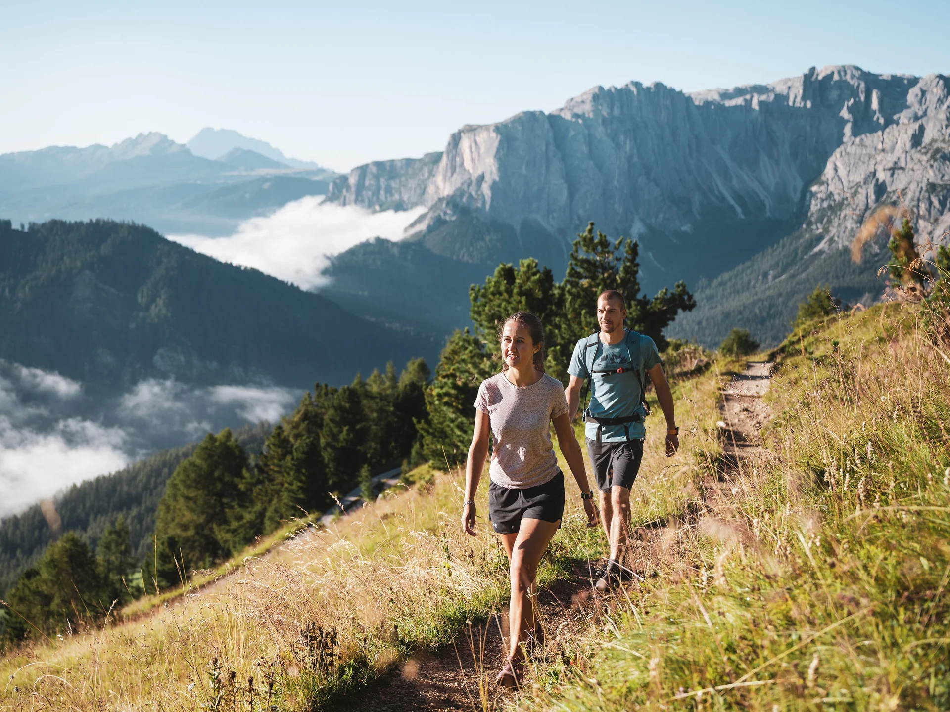 Zwei Wanderer gehen auf einem Bergpfad vor einer wolkenverhangenen Bergkulisse