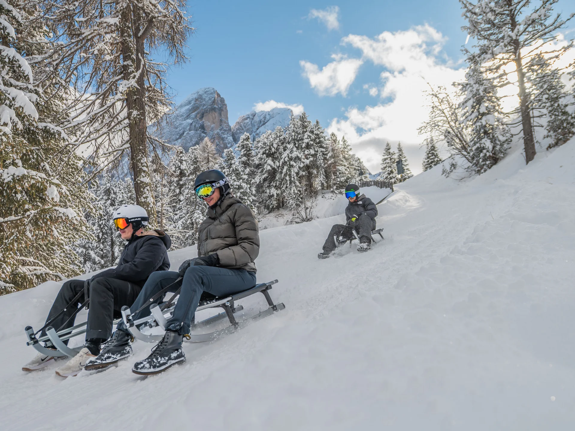 Drei Personen rodeln auf einer verschneiten Piste durch den winterlichen Wald