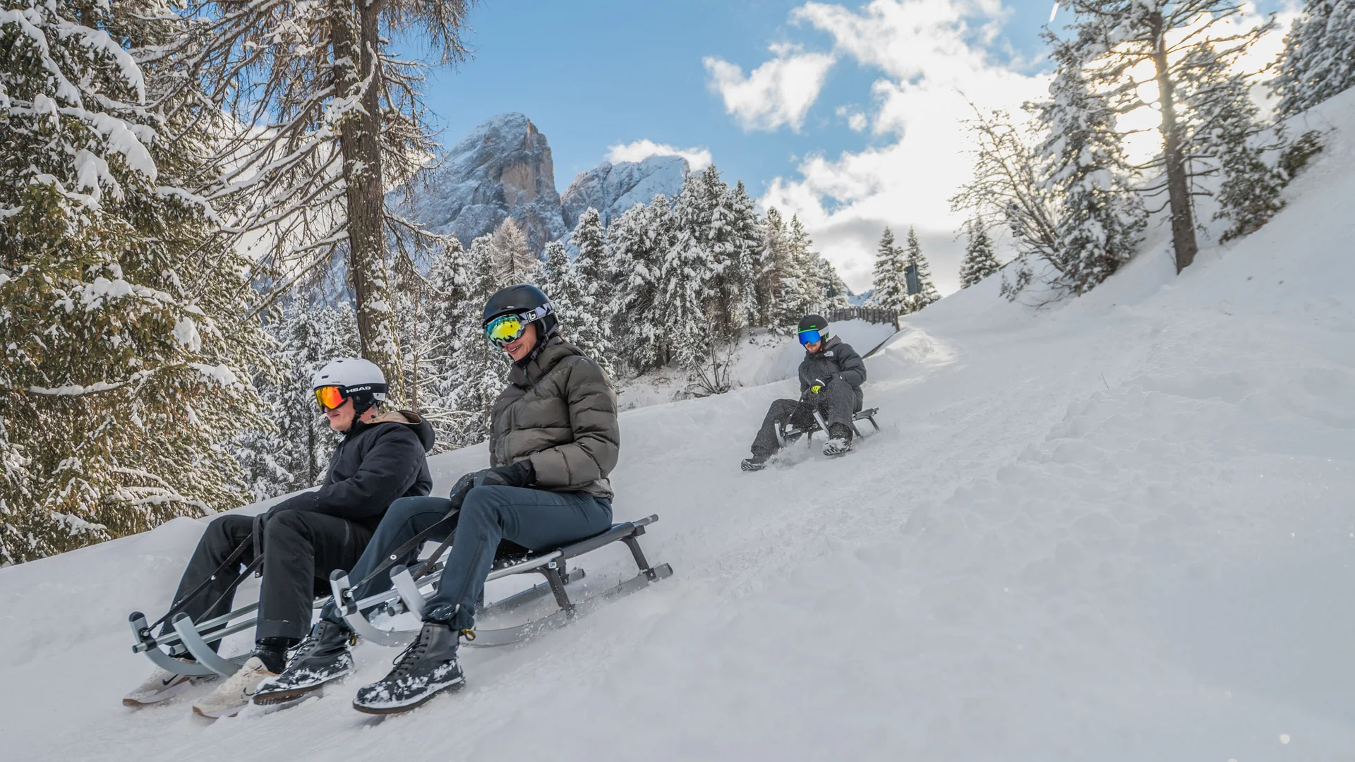 Drei Personen rodeln auf einer verschneiten Piste durch den winterlichen Wald