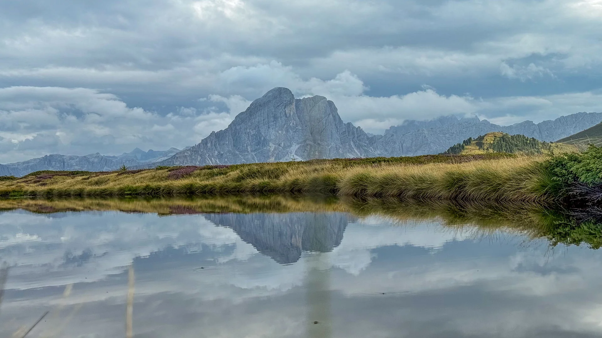 Berggipfel und bewachsene Ufer spiegeln sich in einem stillen See unter bewölktem Himmel