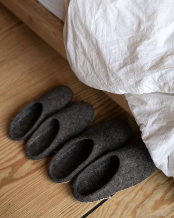 Vier graue Filzpantoffeln neben einem Bett mit weißer Bettwäsche