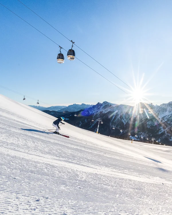 Skifahrer auf einer sonnigen Piste vor schneebedeckten Bergen und Gondeln