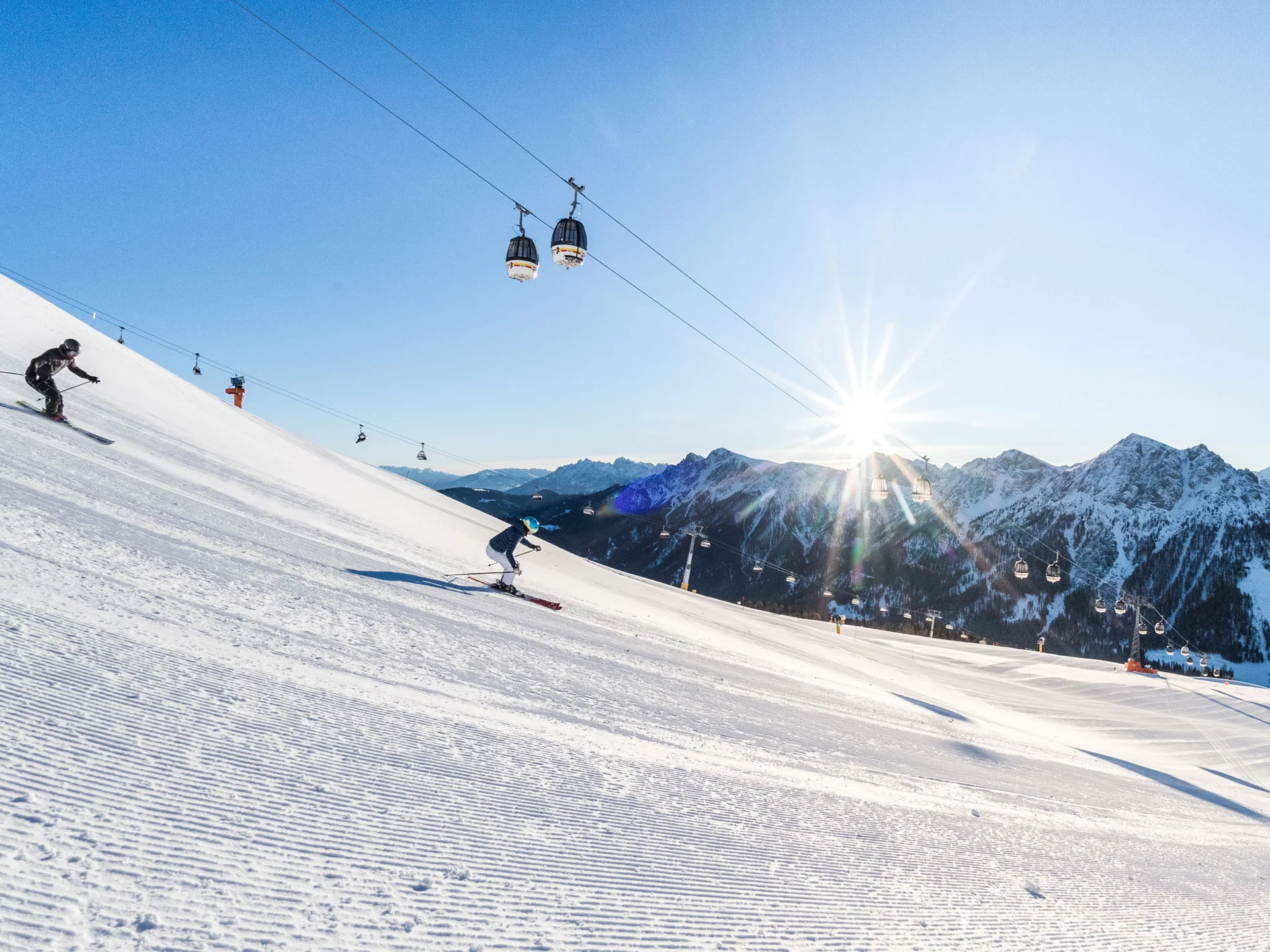 Skifahrer auf einer sonnigen Piste vor schneebedeckten Bergen und Gondeln