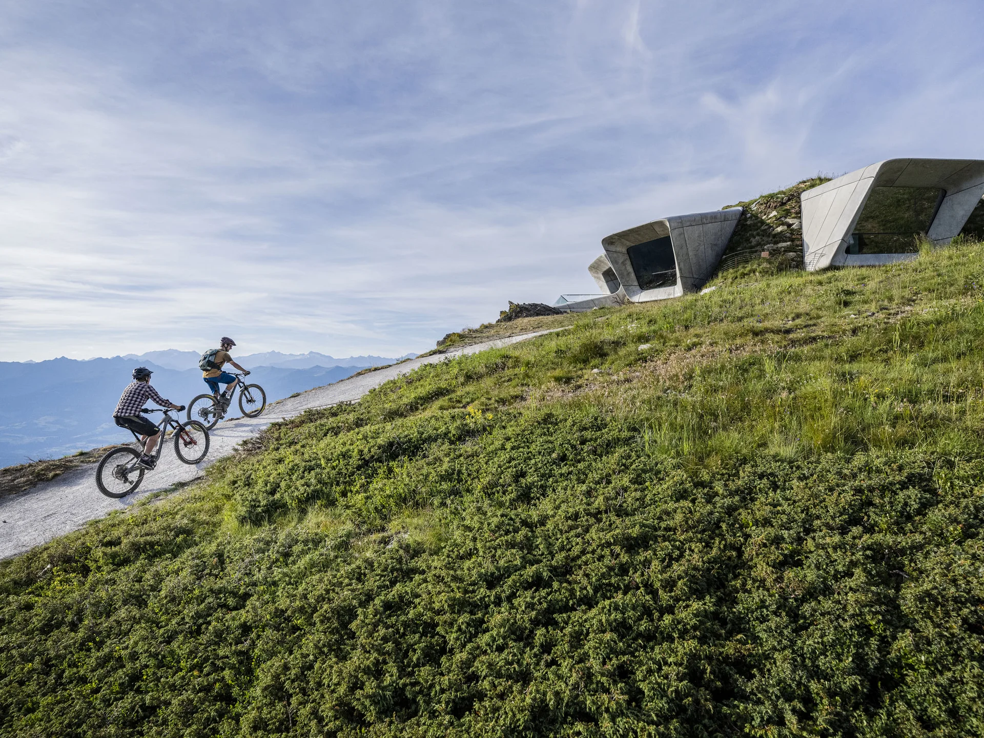 Zwei Mountainbiker fahren auf einem Bergweg an modernen Betonbauten vorbei