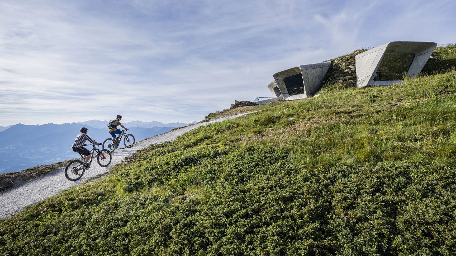 Zwei Mountainbiker fahren auf einem Bergweg an modernen Betonbauten vorbei
