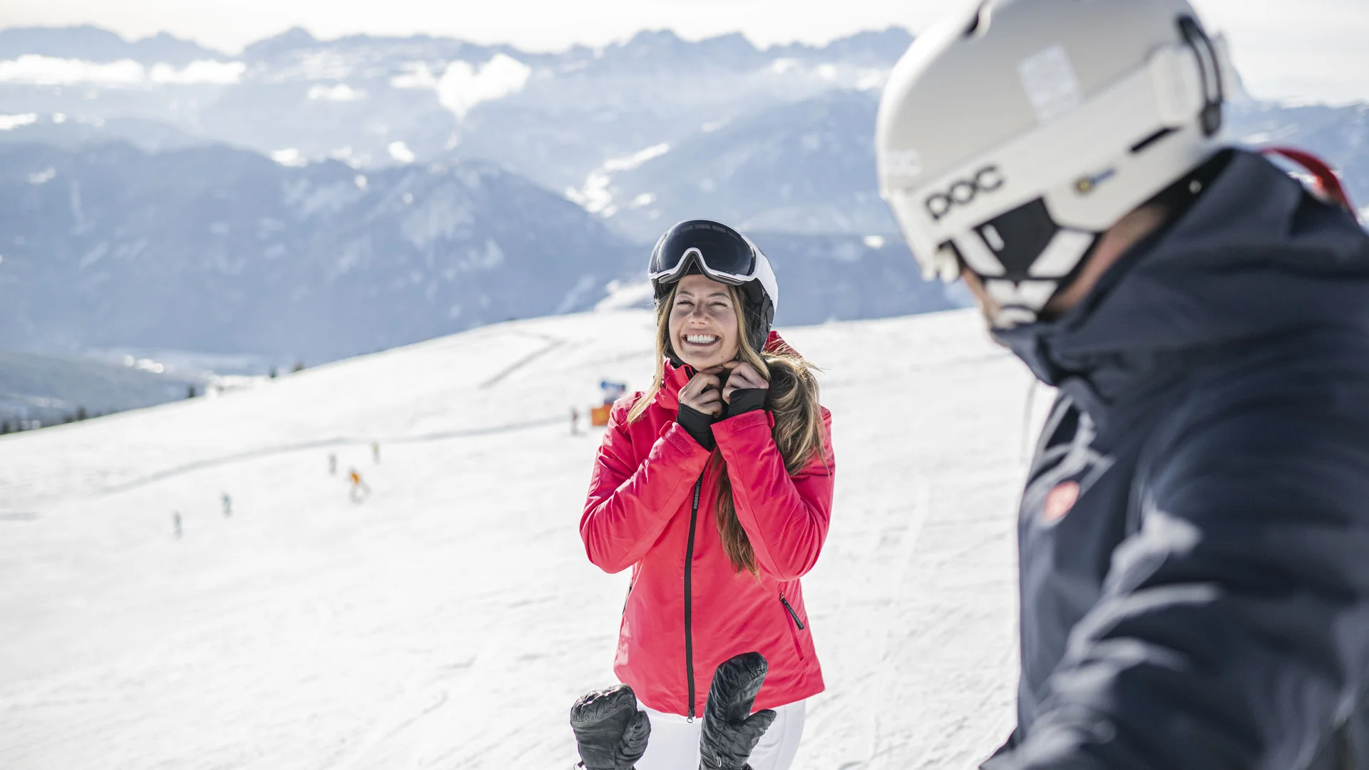 Naturparks Fanes-Sennes-Prags und Puez-Geisler | Chalet Löm Lächelnde Skifahrerin in roter Jacke auf einer verschneiten Piste, ein weiterer Skifahrer im Vordergrund