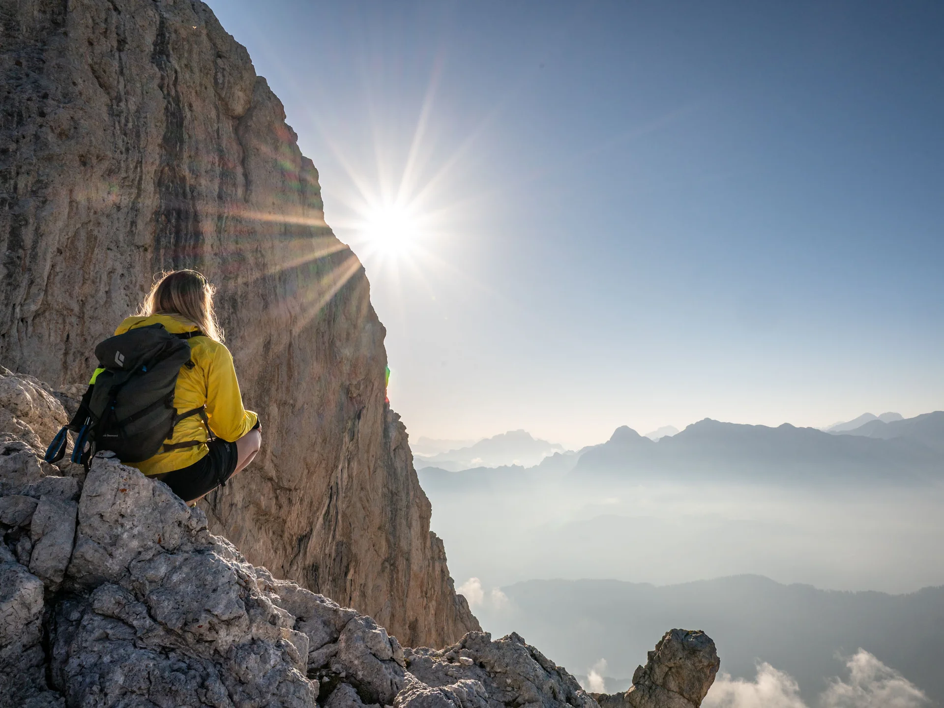 Naturparks Fanes-Sennes-Prags und Puez-Geisler | Chalet Löm Wanderin mit gelber Jacke und Rucksack sitzt an einer Felskante mit Blick auf sonnenbeschienene Berge