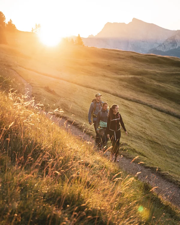 Naturparks Fanes-Sennes-Prags und Puez-Geisler | Chalet Löm Drei Wanderer gehen bei Sonnenuntergang auf einem Bergpfad durch goldenes Gras