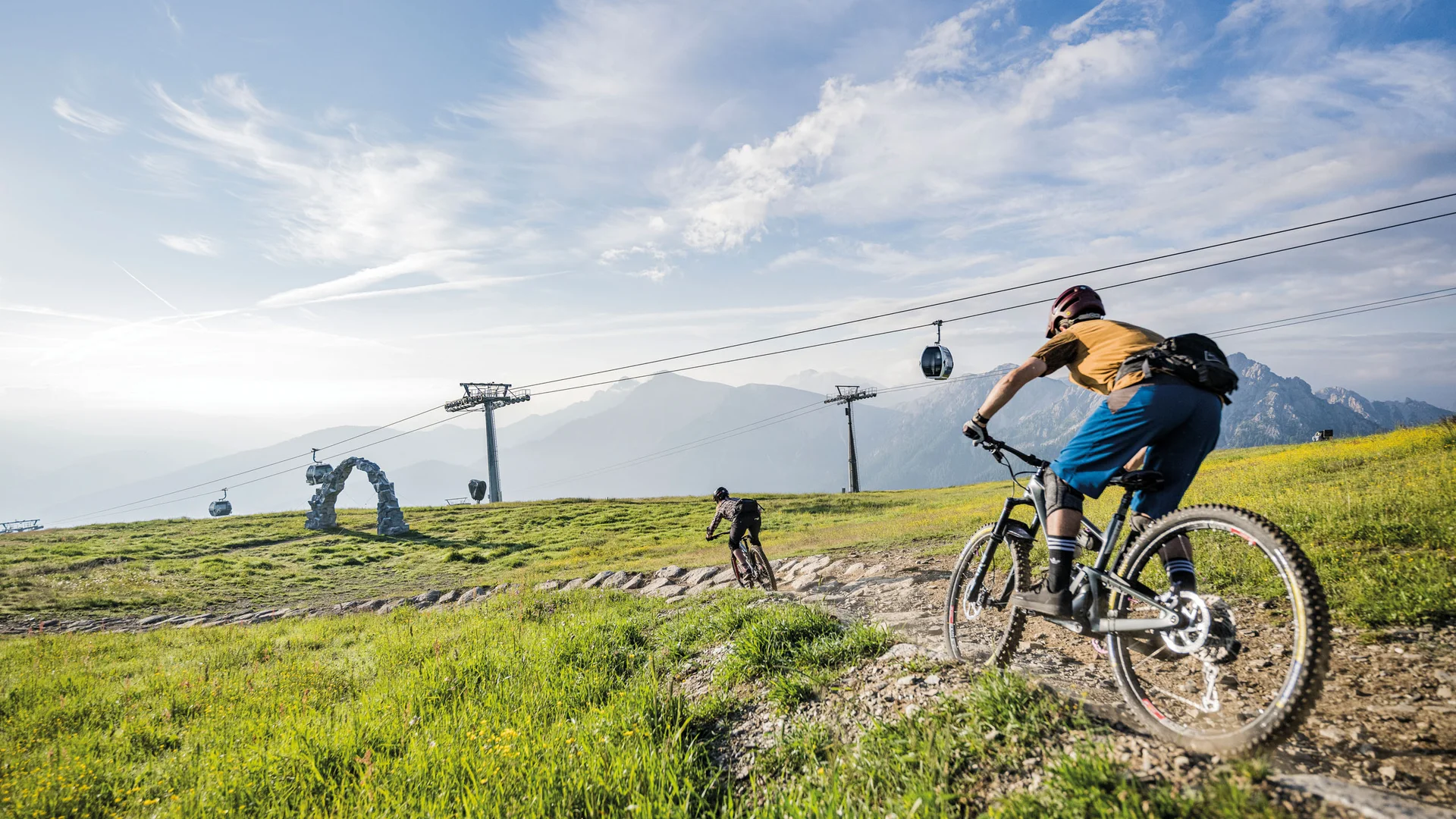 Zwei Mountainbiker fahren über einen Bergpfad, mit Seilbahn und Gipfeln im Hintergrund