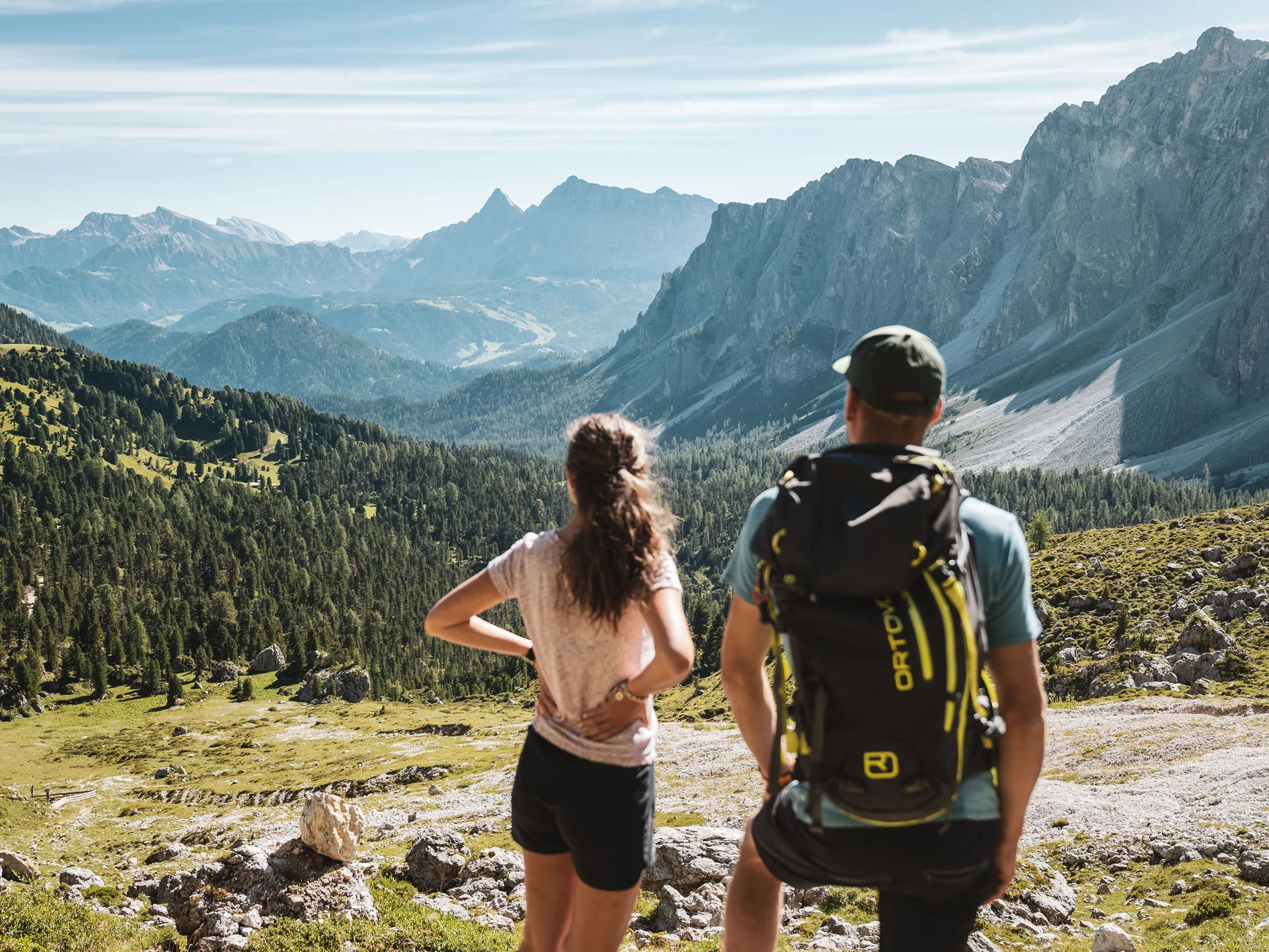 Zwei Wanderer blicken auf ein bewaldetes Alpental und die umliegenden Berge