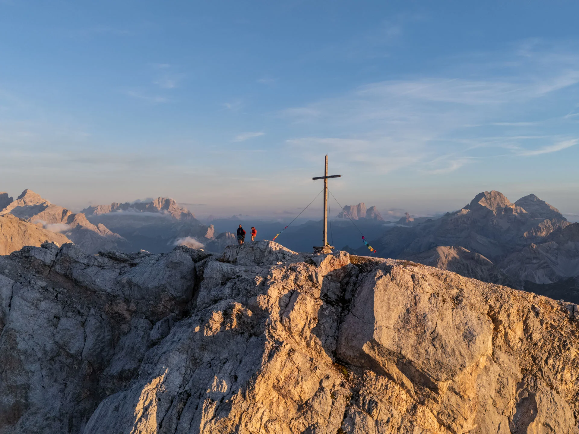 Naturparks Fanes-Sennes-Prags und Puez-Geisler | Chalet Löm Zwei Wanderer stehen neben einem Gipfelkreuz auf einem felsigen Berggipfel mit Alpenpanorama