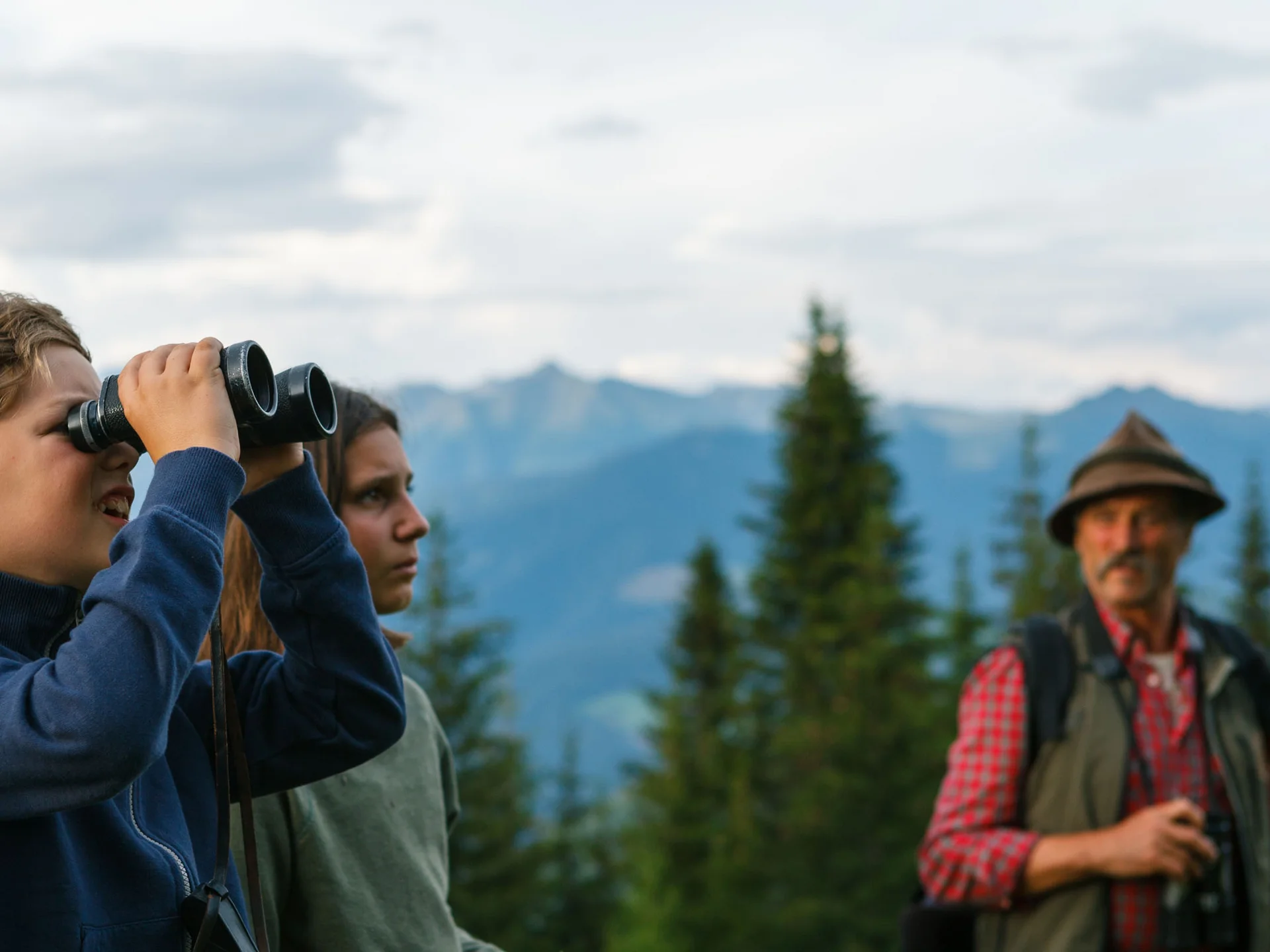 Zwei Jugendliche beobachten mit einem Fernglas die Berglandschaft, während ein Mann danebensteht