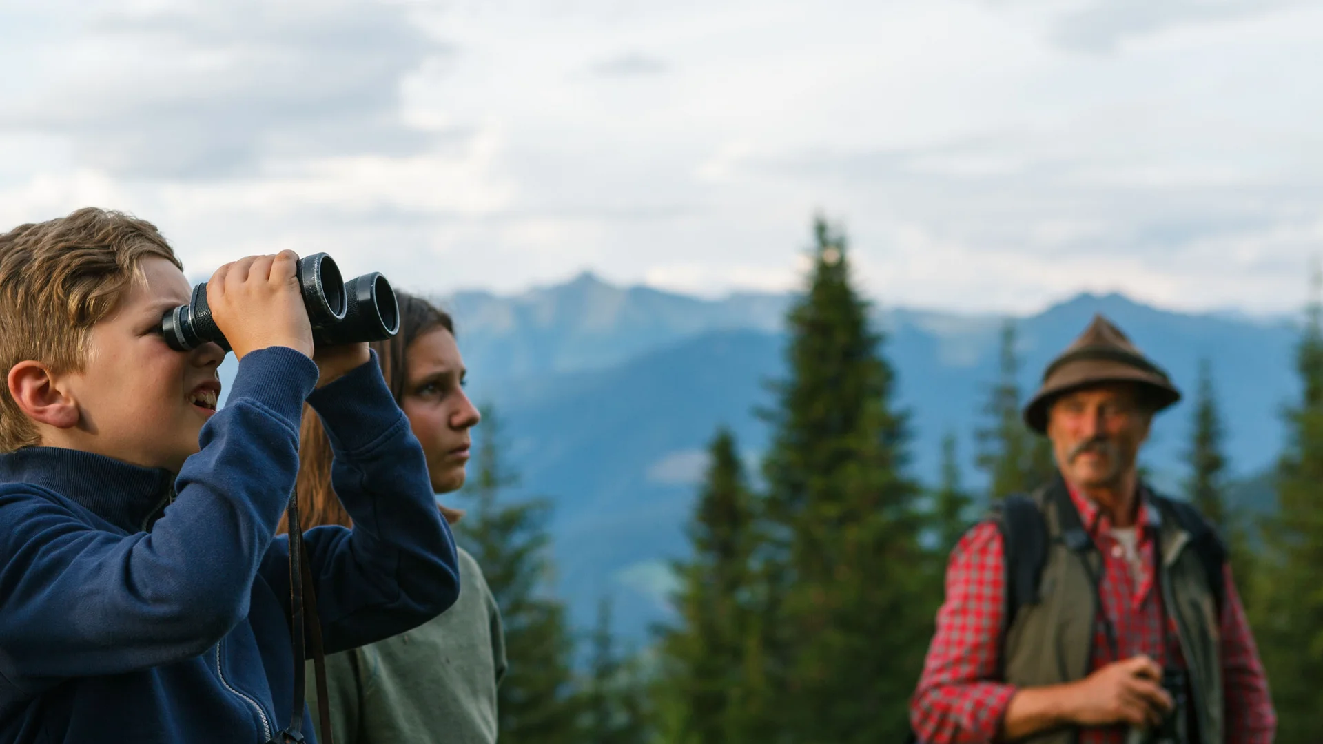 Zwei Jugendliche beobachten mit einem Fernglas die Berglandschaft, während ein Mann danebensteht