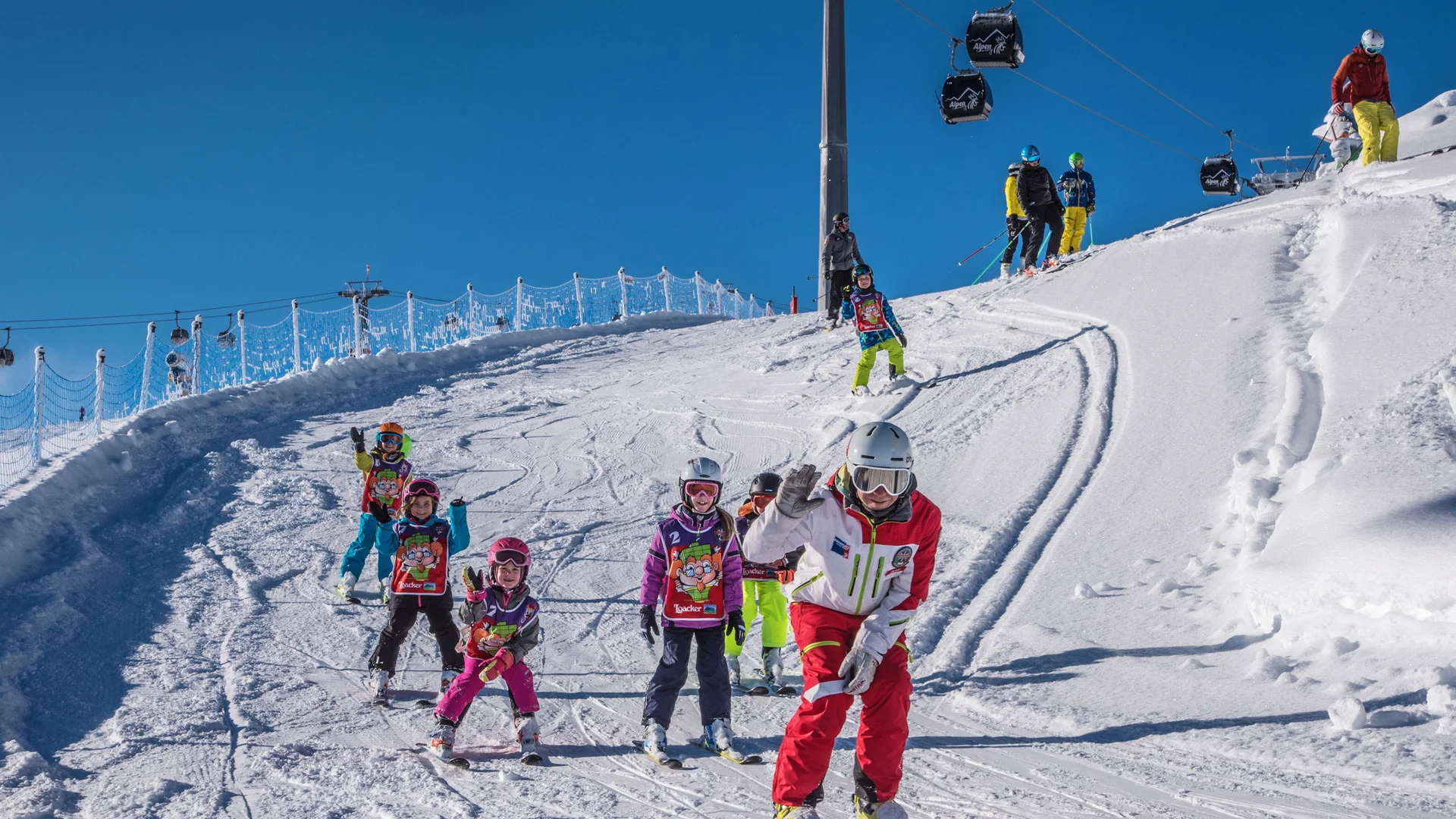 Skifahrer und Kinder fahren gemeinsam eine verschneite Piste hinunter, Gondeln schweben darüber