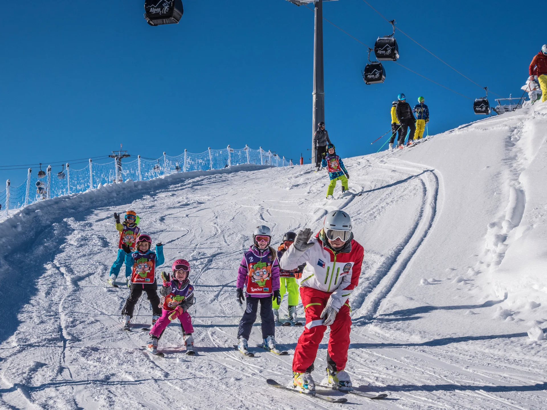 Skifahrer und Kinder fahren gemeinsam eine verschneite Piste hinunter, Gondeln schweben darüber