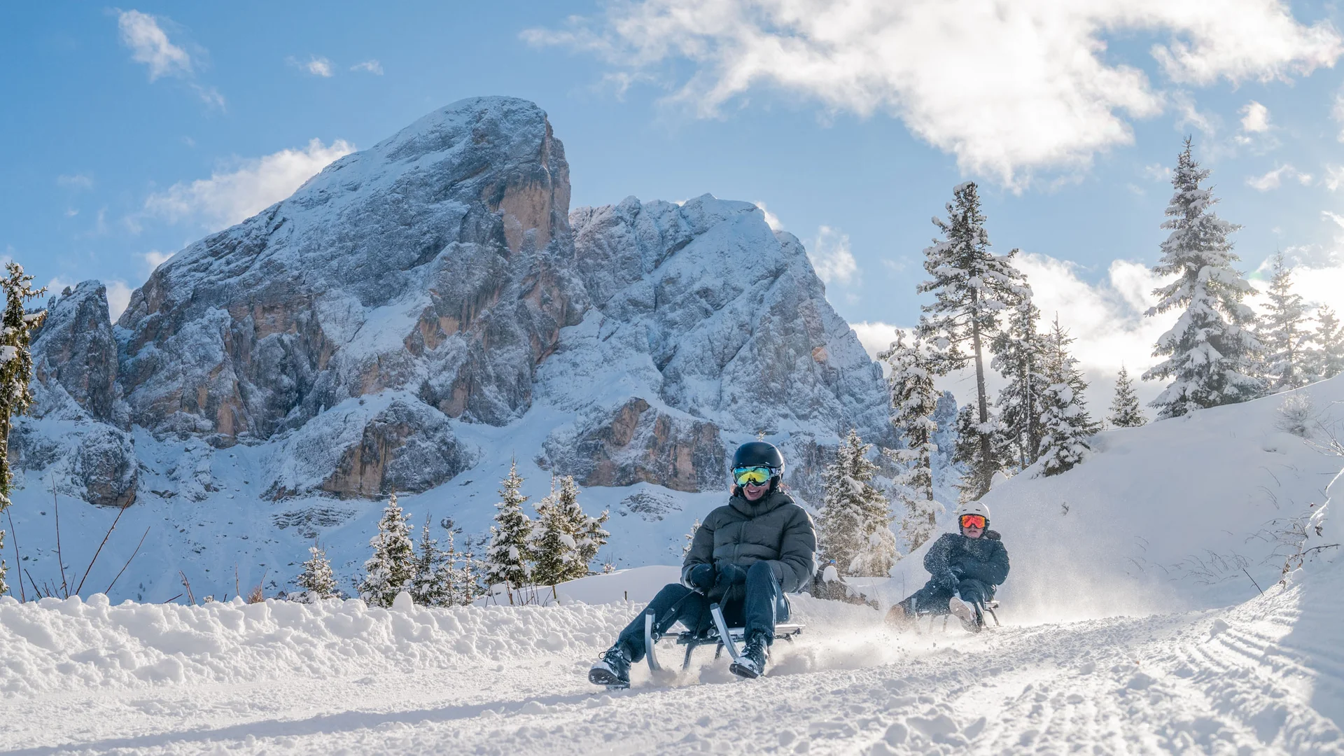 Zwei Personen rodeln vor einer verschneiten Bergkulisse durch den Wald