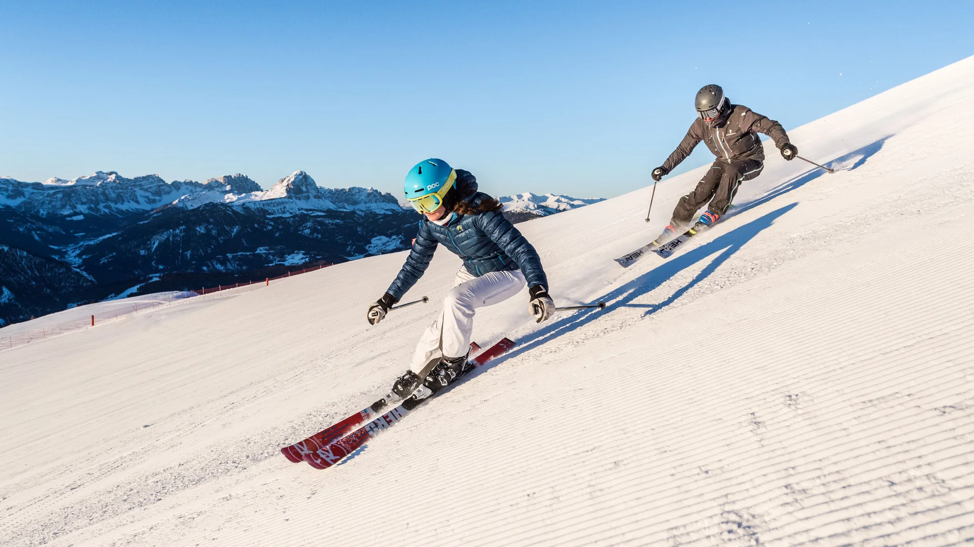 Zwei Skifahrer fahren auf einer präparierten Piste vor schneebedeckten Bergen