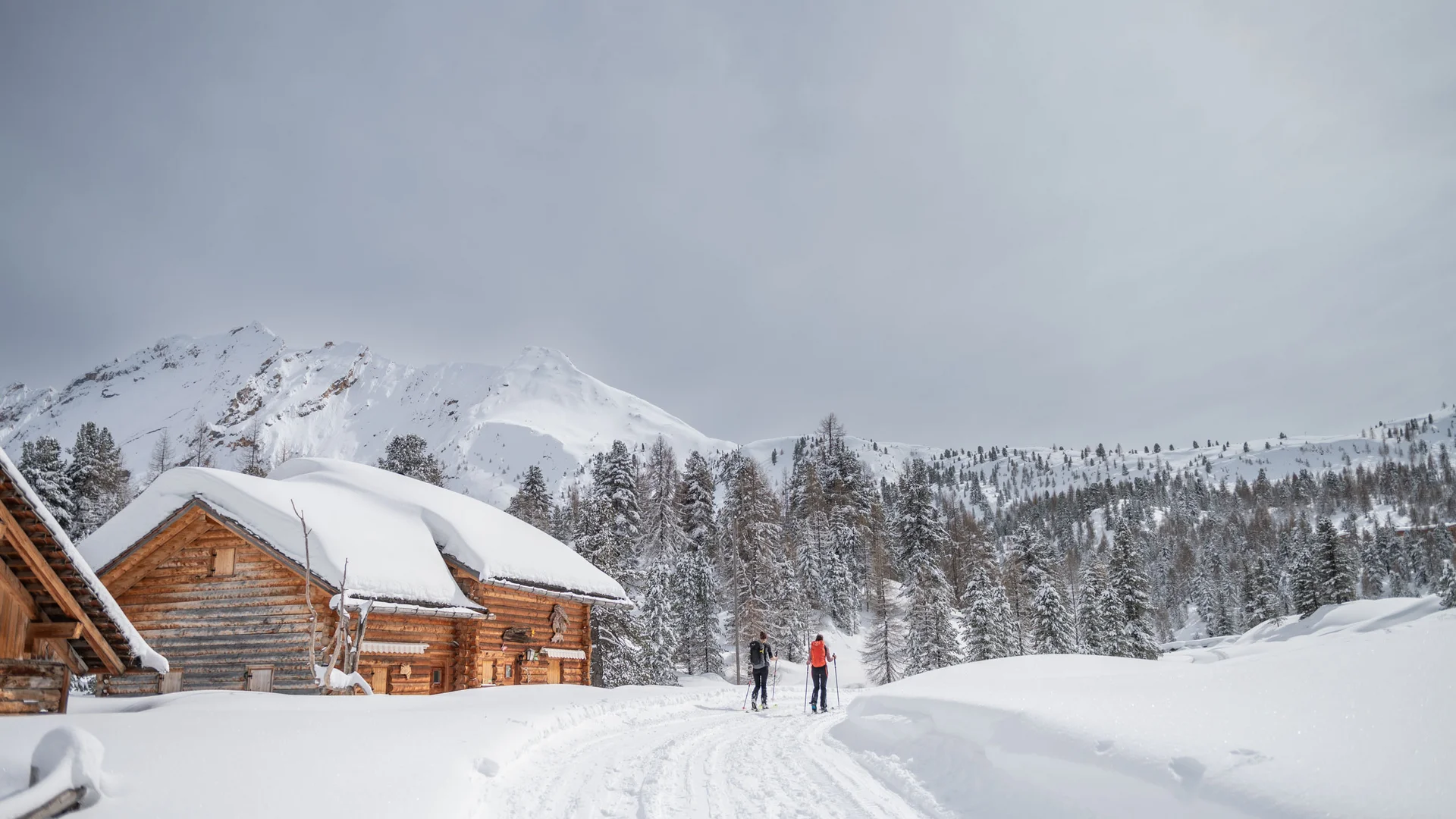 Zwei Skilangläufer auf einem verschneiten Weg zwischen Berghütten und schneebedeckten Bergen