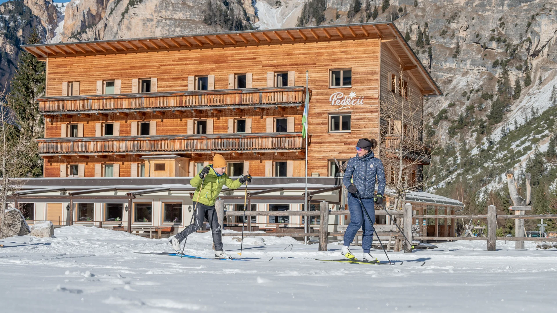 Zwei Langläufer vor einem Holzhotel in verschneiter Berglandschaft