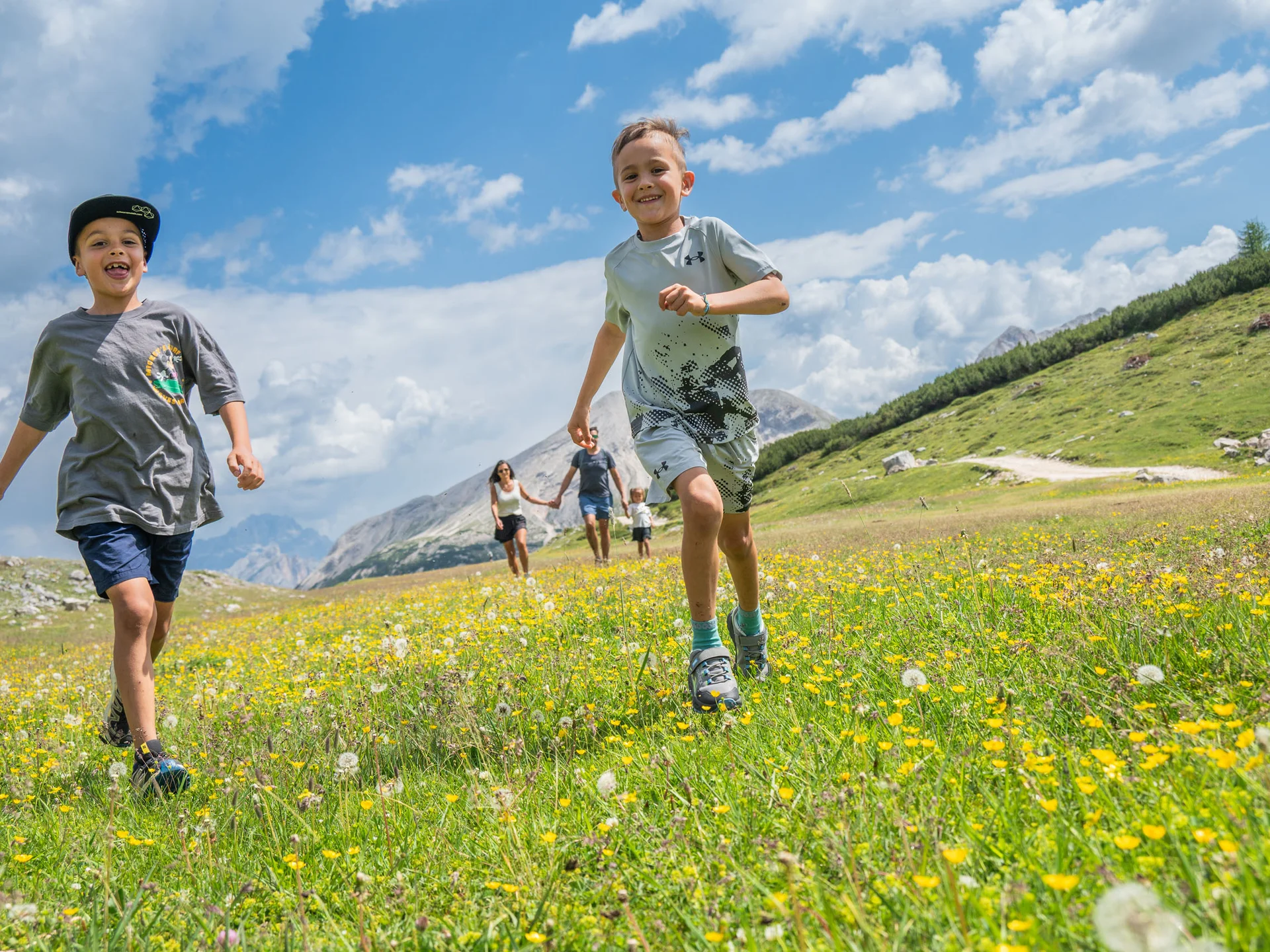 Naturparks Fanes-Sennes-Prags und Puez-Geisler | Chalet Löm Kinder laufen über eine blühende Bergwiese, dahinter eine Familie und alpine Gipfel