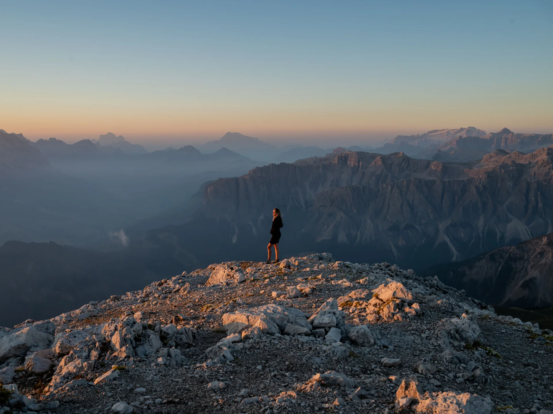 Naturparks Fanes-Sennes-Prags und Puez-Geisler | Chalet Löm Frau steht auf einem felsigen Berggipfel vor einer weitläufigen Bergkulisse im Abendlicht