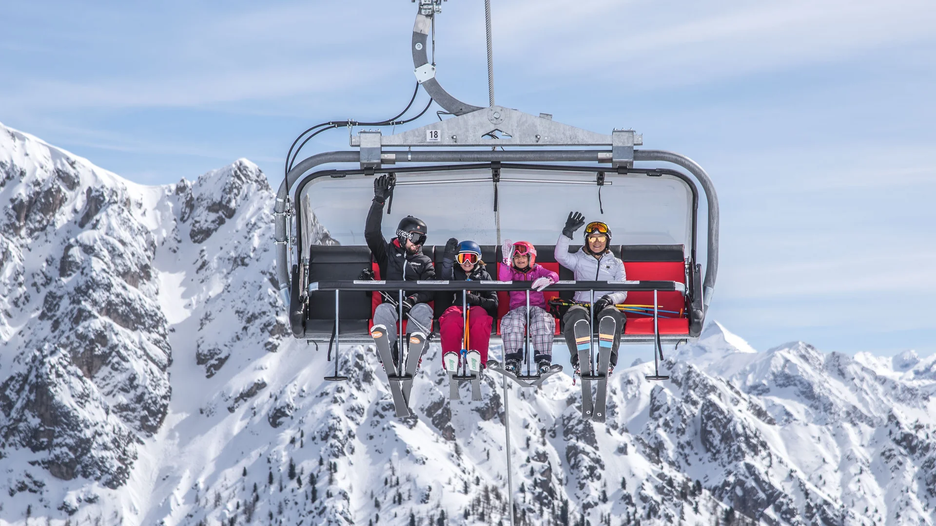 Vier Skifahrer winken auf einem Sessellift vor schneebedeckten Bergen