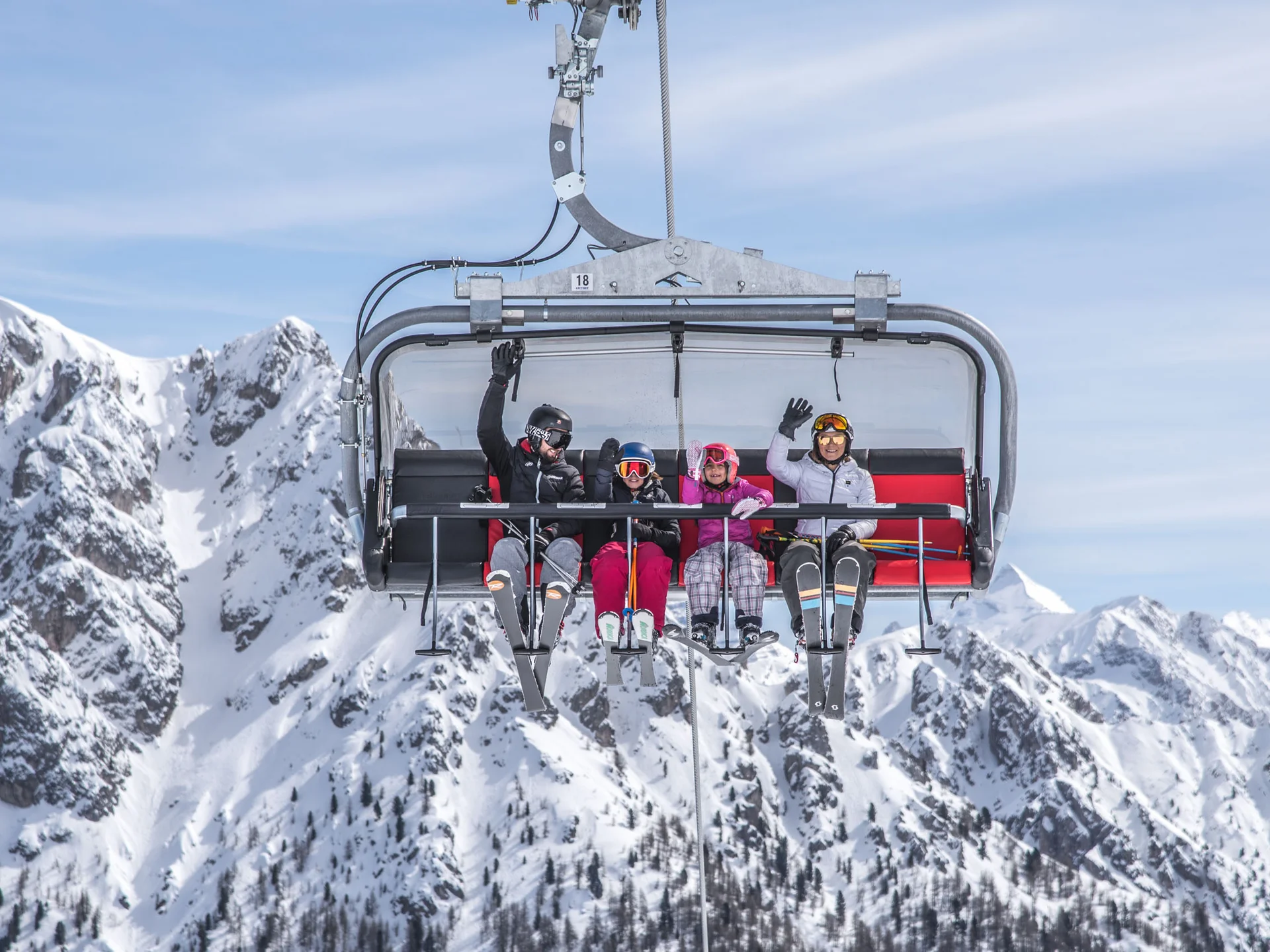 Naturparks Fanes-Sennes-Prags und Puez-Geisler | Chalet Löm Vier Skifahrer winken auf einem Sessellift vor schneebedeckten Bergen