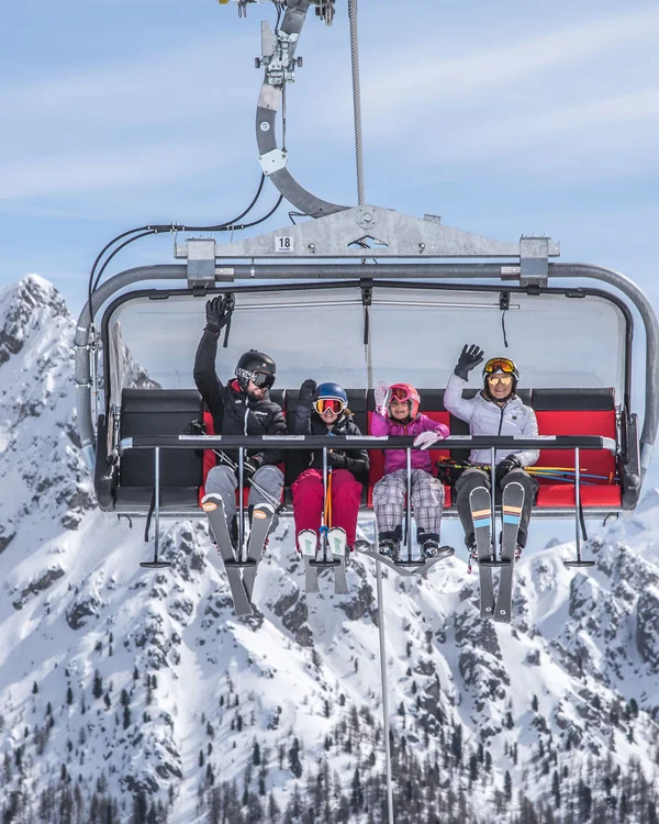 Vier Skifahrer winken auf einem Sessellift vor schneebedeckten Bergen
