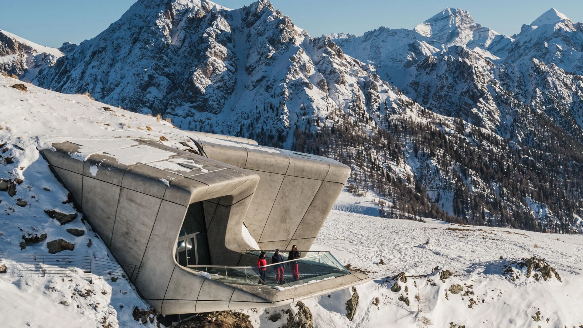 Modernes Gebäude mit Aussichtsplattform in verschneiter Berglandschaft, drei Menschen am Geländer