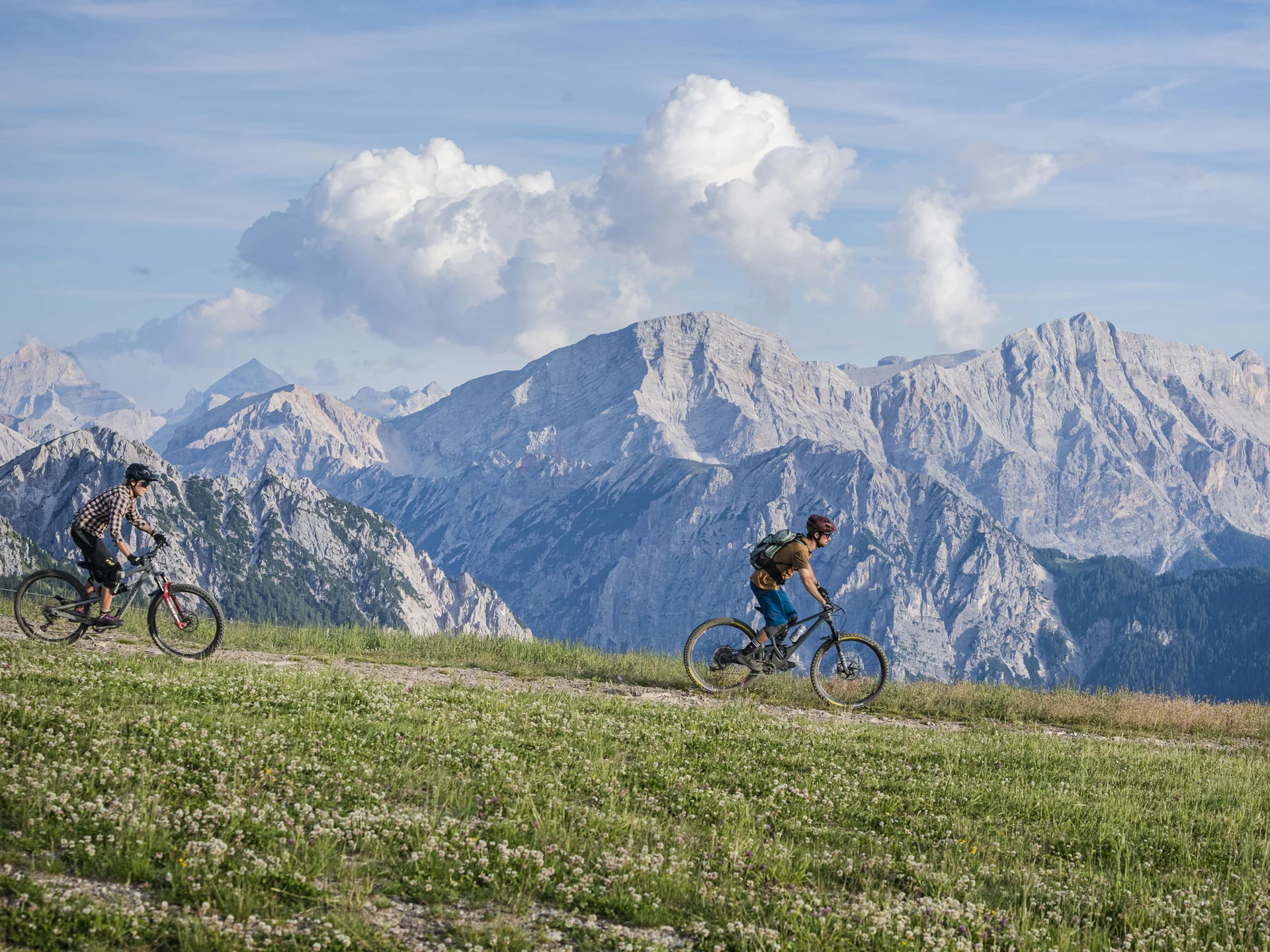 Zwei Mountainbiker fahren über einen Wiesenweg vor einer Kulisse aus schroffen Bergen und Wolken