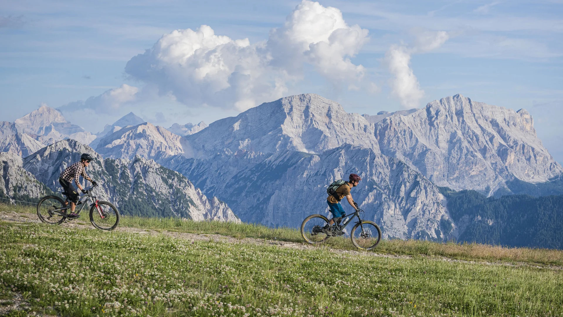 Zwei Mountainbiker fahren über einen Wiesenweg vor einer Kulisse aus schroffen Bergen und Wolken