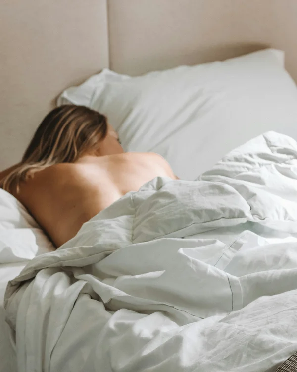 Frau liegt mit dem Rücken zur Kamera in einem Bett mit weißer Bettwäsche